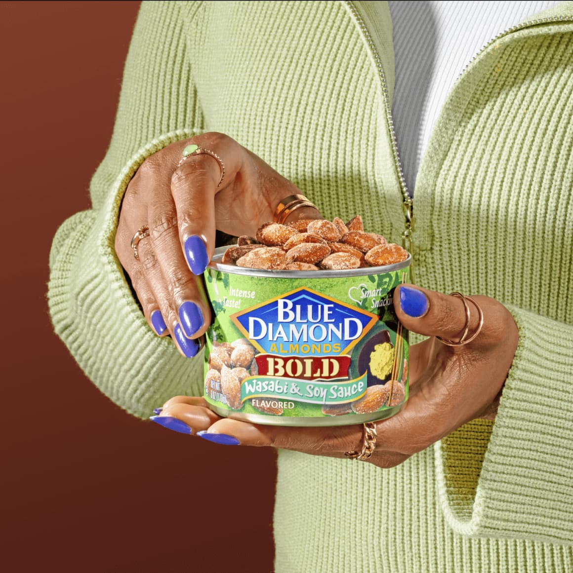 A woman wearing a green sweater holding a can of Wasabi & Soy Sauce flavored almonds in her hands.