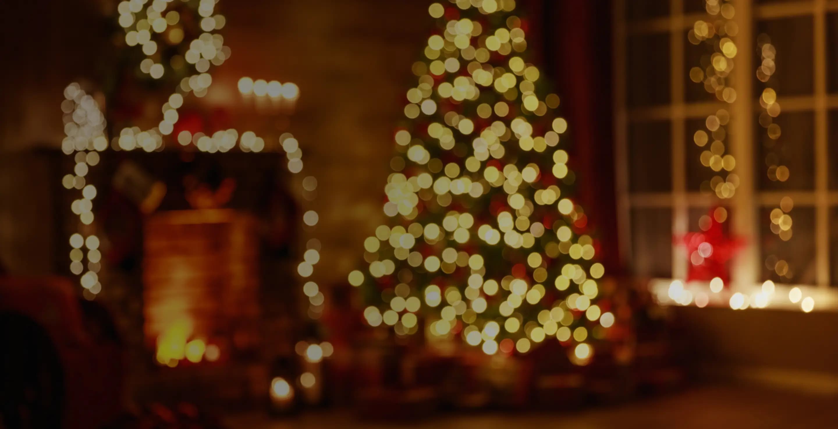 A warmly lit, softly blurred holiday scene featuring a glowing Christmas tree decorated with string lights. In the background, a fireplace and window are also adorned with festive lights, creating a cozy, golden bokeh effect throughout the room.