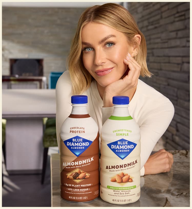 A woman with short blonde hair smiles next to two cartons of Blue Diamond Almondmilk, one chocolate protein and one unsweetened simple, on a table indoors.