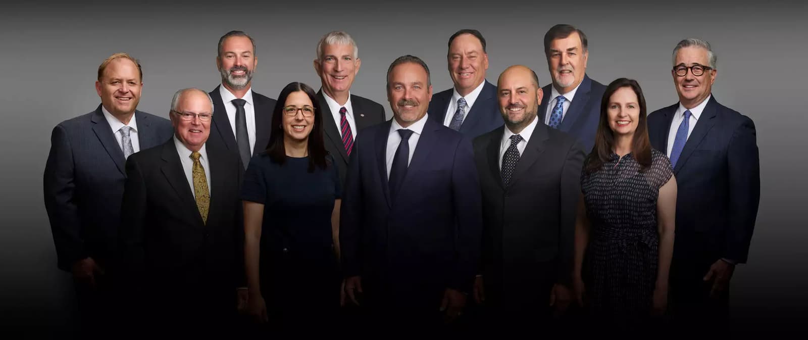 Twelve professionally dressed people, both men and women, stand together in two rows, posing and smiling against a dark gradient background.