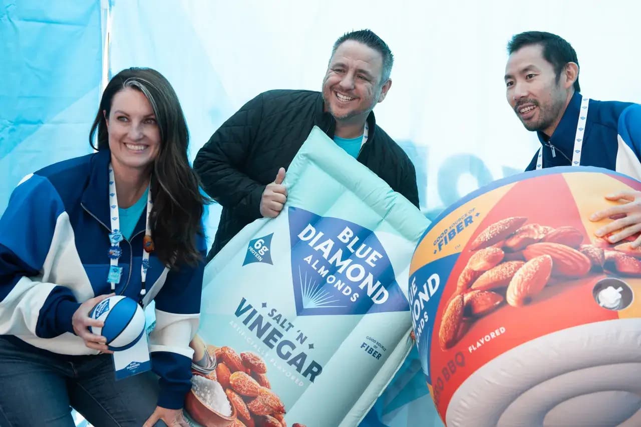 Three people smile while posing with oversized inflatable Blue Diamond almond snack bags and a small blue-and-white basketball.