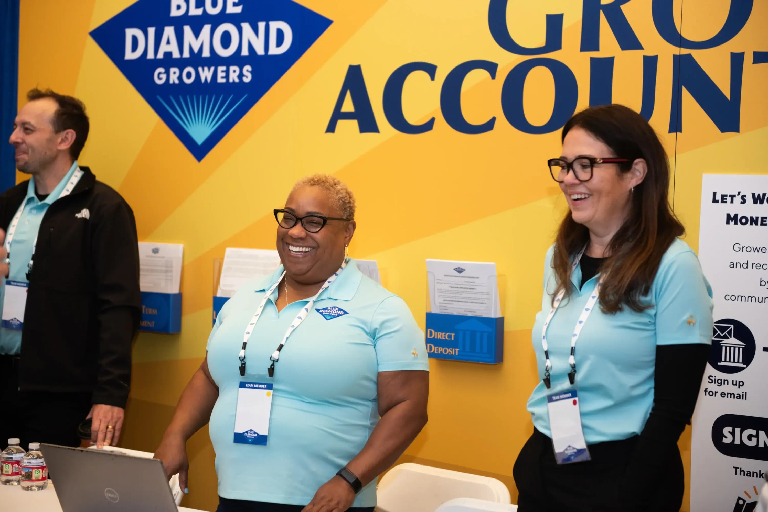Three people wearing conference badges stand and smile at a Blue Diamond Growers booth with yellow and blue signage in the background.