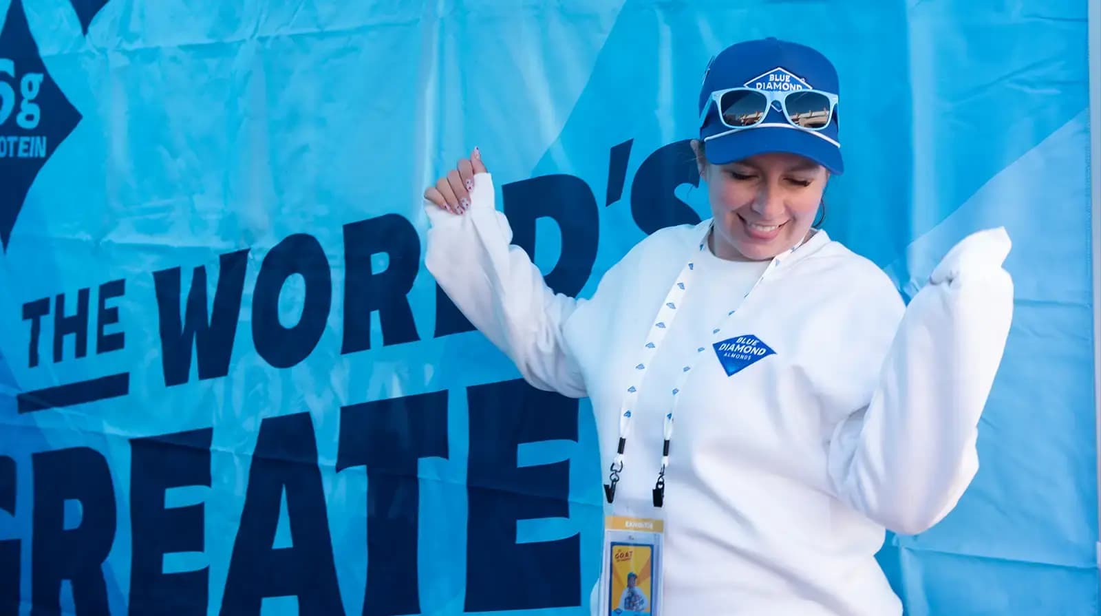 Person in a white sweatshirt and blue cap stands in front of a blue banner that says THE WORLDS GREATEST, smiling and raising both arms.