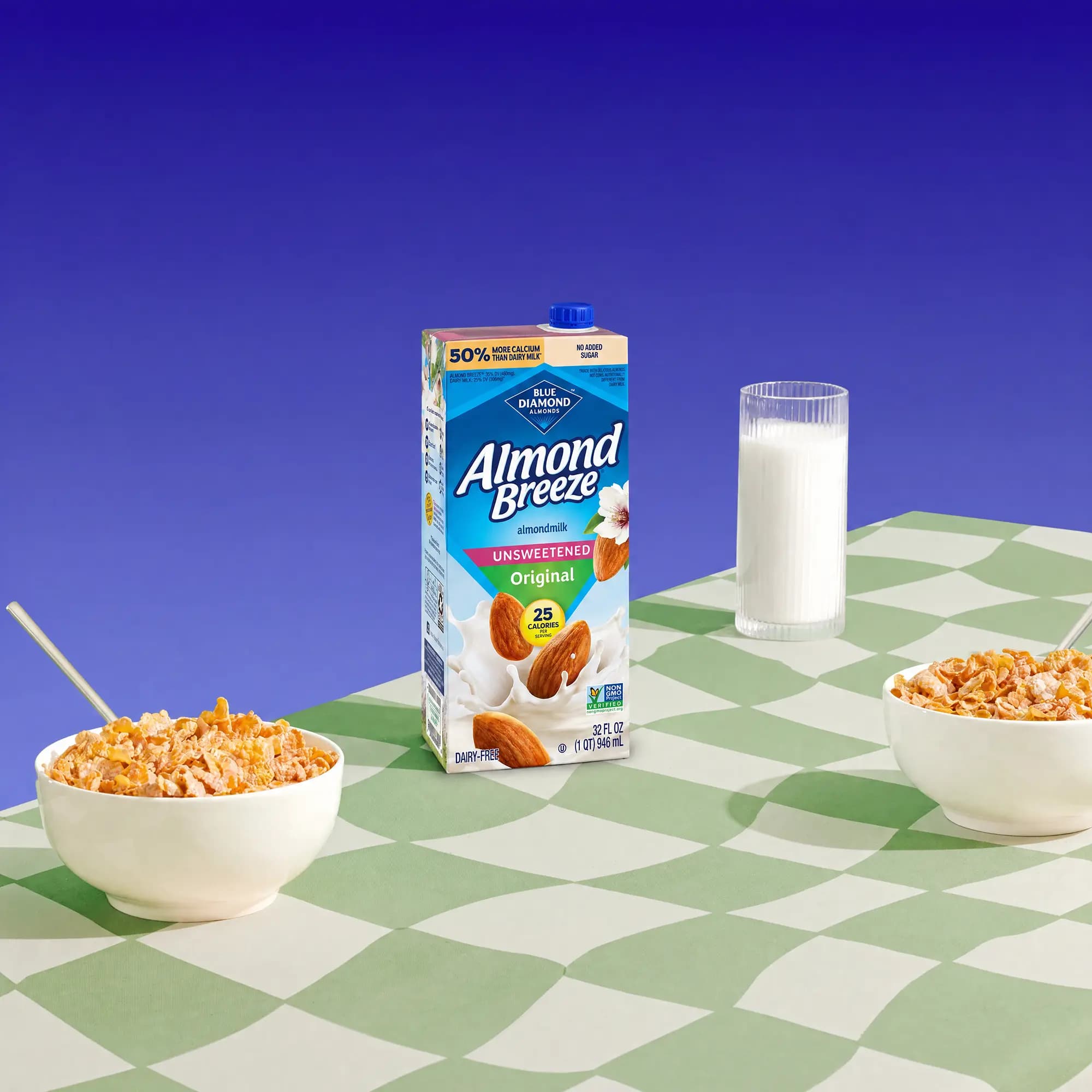 Carton of Almond Breeze Unsweetened Original almondmilk displayed on a green-and-white checkered table with a glass of almondmilk and two bowls of cereal against a vibrant blue background.