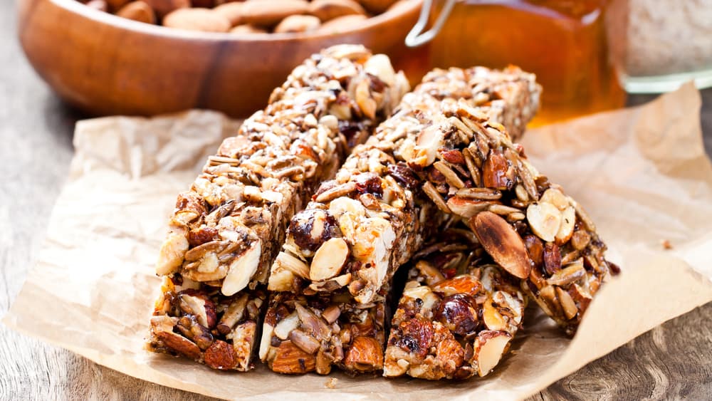Stack of homemade almond, seed, and dried fruit granola bars on parchment paper, with a bowl of almonds and a jar of honey softly blurred in the background.