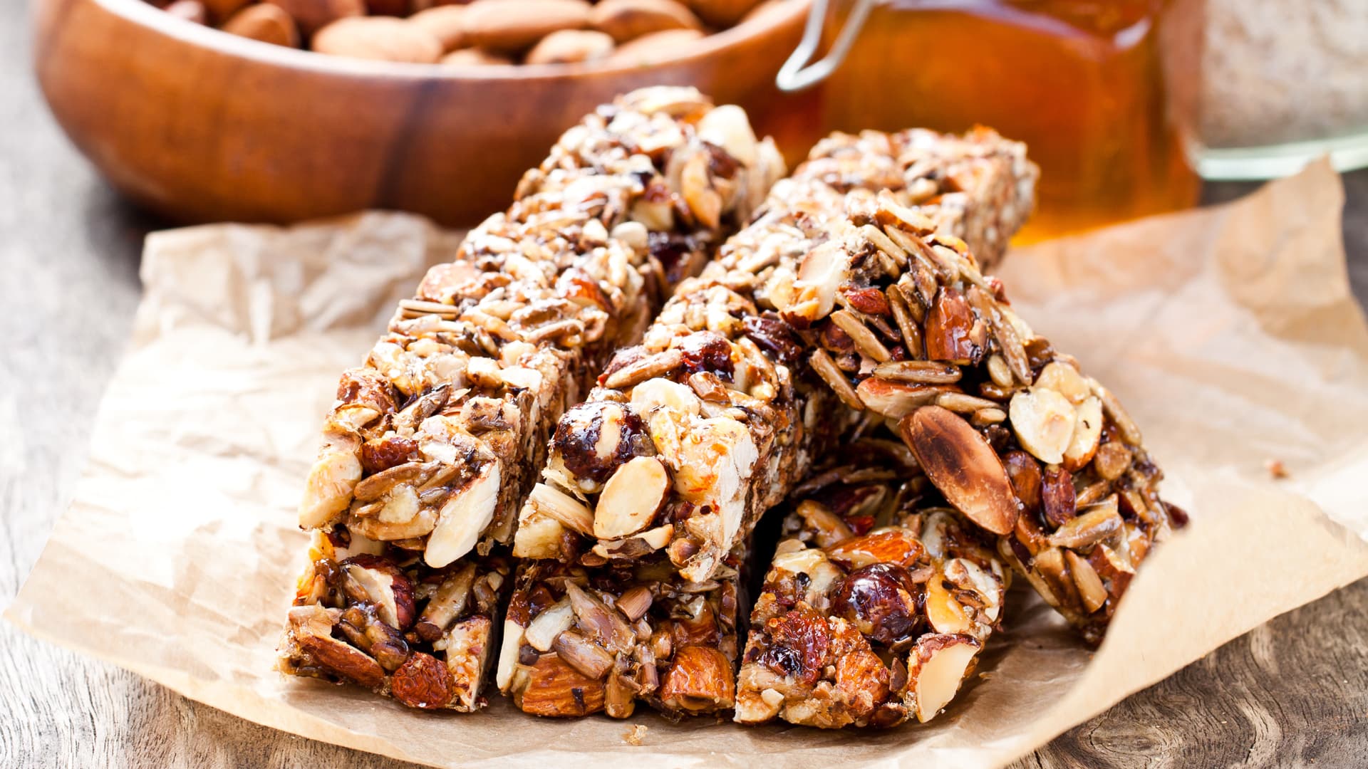 Stack of homemade almond, seed, and dried fruit granola bars on parchment paper, with a bowl of almonds and a jar of honey softly blurred in the background.