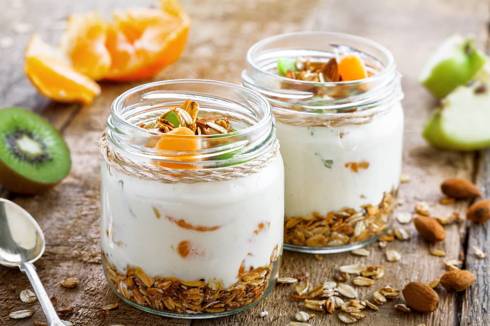 Two glass jars of creamy yogurt layered with granola and topped with sliced kiwi, mandarin orange segments, almonds, and pumpkin seeds on a rustic wooden table, with fresh fruit and scattered oats in the background.