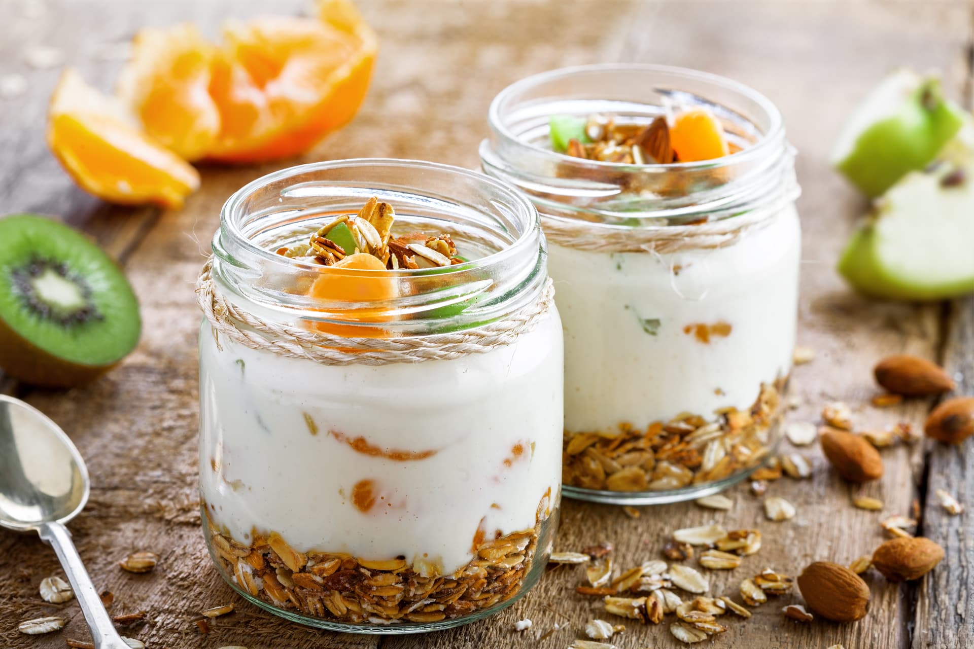 Two glass jars of creamy yogurt layered with granola and topped with sliced kiwi, mandarin orange segments, almonds, and pumpkin seeds on a rustic wooden table, with fresh fruit and scattered oats in the background.