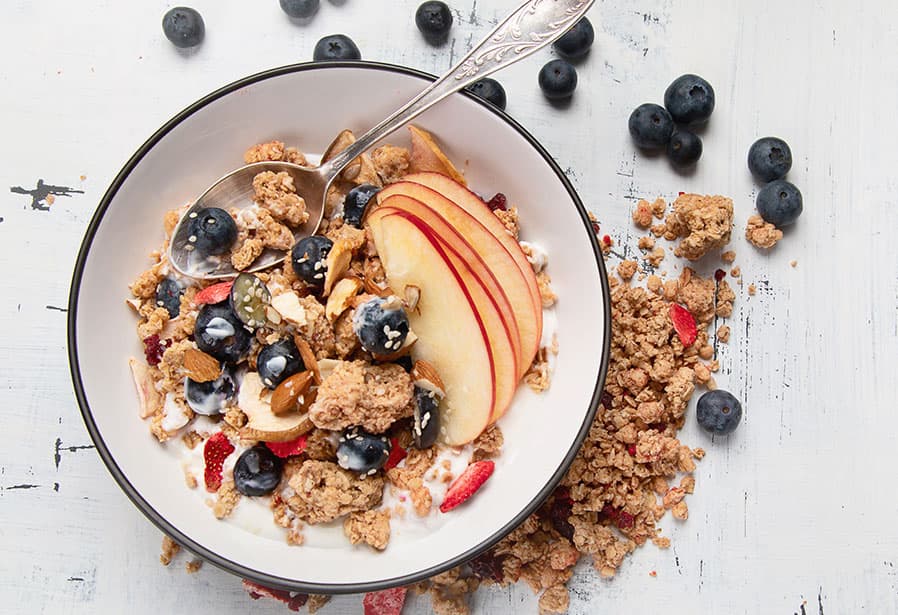 Overhead view of a bowl of yogurt topped with granola, fresh blueberries, sliced apple, almonds, and seeds, with a spoon resting in the bowl and scattered blueberries and granola on a white wooden surface.