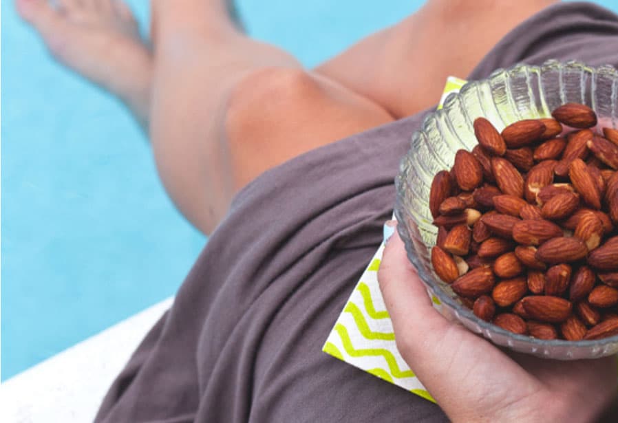 Person relaxing poolside while holding a glass bowl filled with whole almonds, with the blue water and outstretched legs softly blurred in the background.