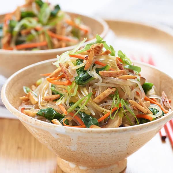 Bowl of Asian-style glass noodles with carrots, leafy greens, scallions, and sliced almonds, garnished with fresh herbs.