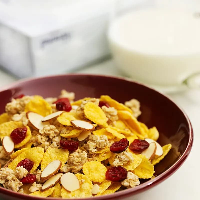 Bowl of cereal with granola, sliced almonds, and dried cranberries served with milk in the background.