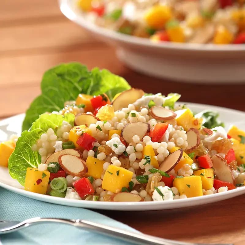 Pearl couscous salad with mango, sliced almonds, red bell pepper, raisins, feta cheese, and fresh herbs on a white plate.
