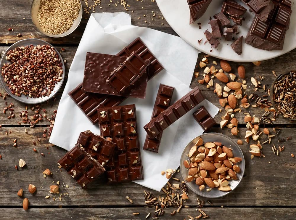 Overhead view of assorted dark chocolate bars on parchment paper surrounded by whole and chopped almonds, grains, and seeds on a rustic wooden table.