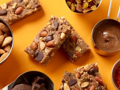 Overhead view of almond snack bars with whole almonds, chocolate chips, and chopped nuts, surrounded by bowls of almonds, raisins, chocolate, almond butter, and spices on a bright orange background.
