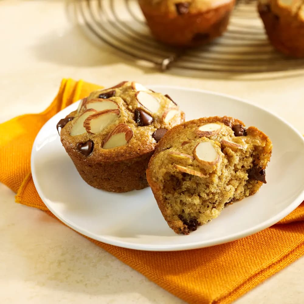 Two almond flour muffins with sliced almonds and chocolate chips on a white plate, with one muffin cut in half to reveal its soft interior.