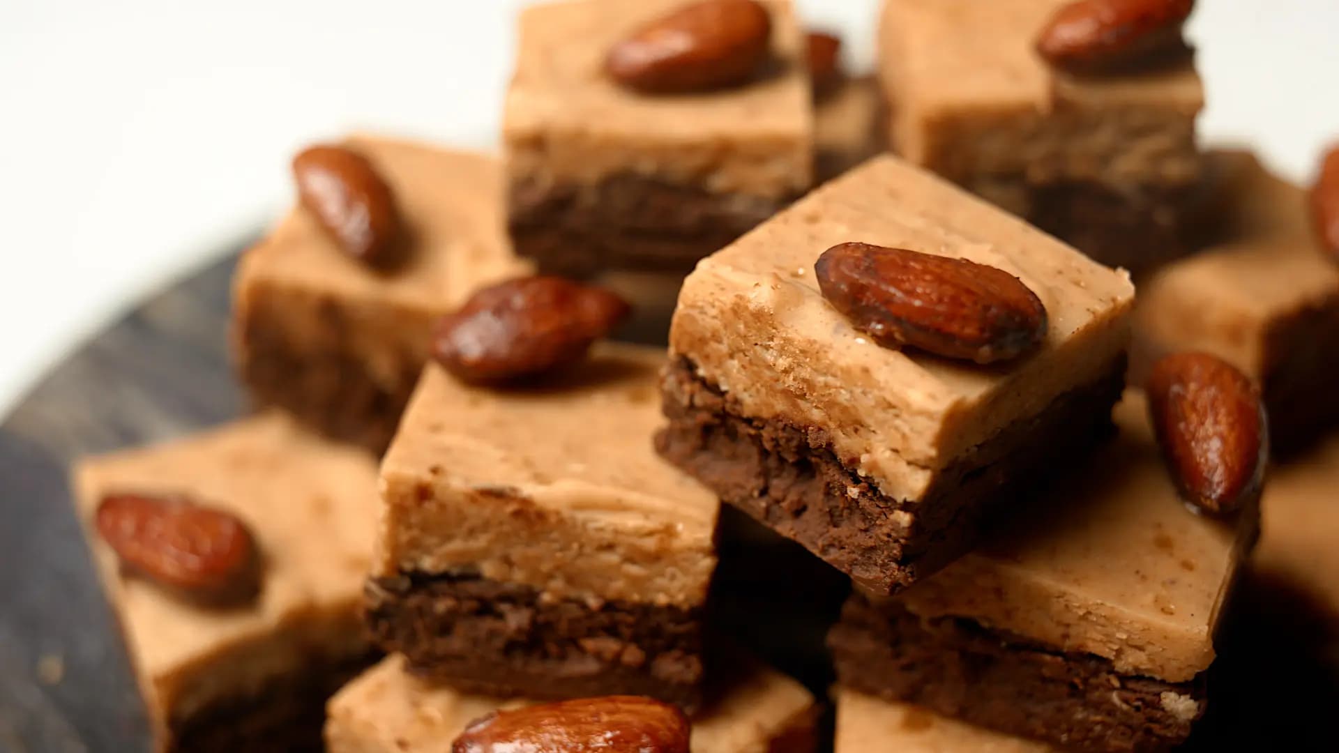 Stack of layered chocolate and almond butter fudge squares topped with whole roasted almonds on a serving plate.