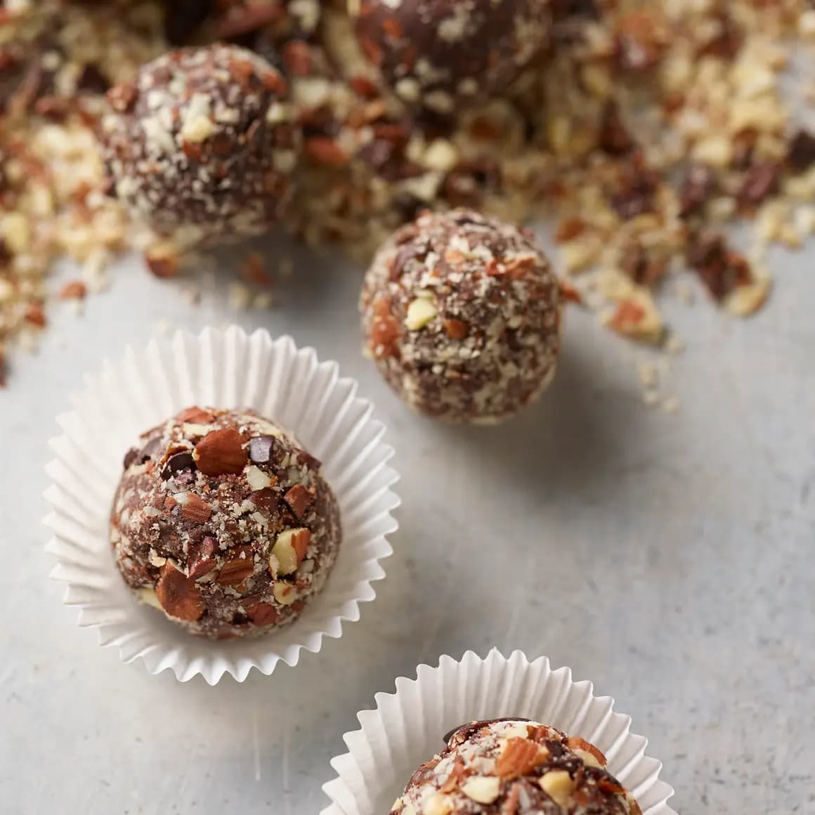 Chocolate almond truffles coated with finely chopped almonds, arranged in white paper cups with additional truffles and chopped almonds in the background.