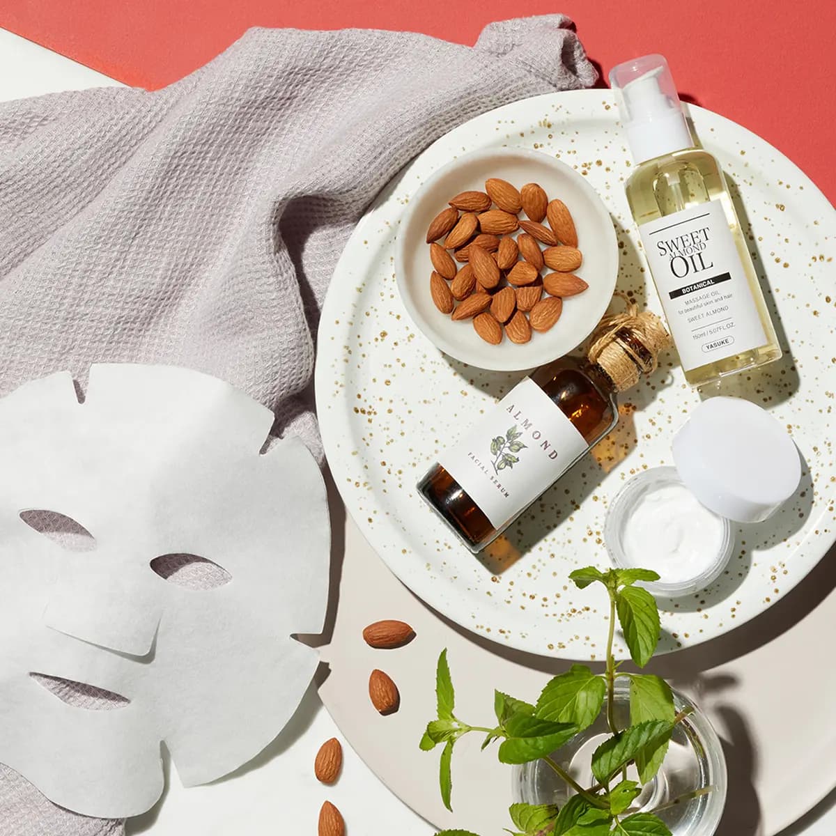 Flat lay of almond-inspired skincare products on a white speckled tray, including bottles of almond oil, a jar of cream, a bowl of whole almonds, a sheet face mask, a soft towel, and fresh mint sprigs on a coral and cream background.