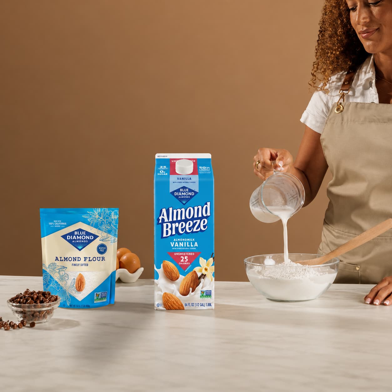 A person pours almond milk into a bowl of flour next to a package of almond flour and a carton of almond milk on a kitchen counter.