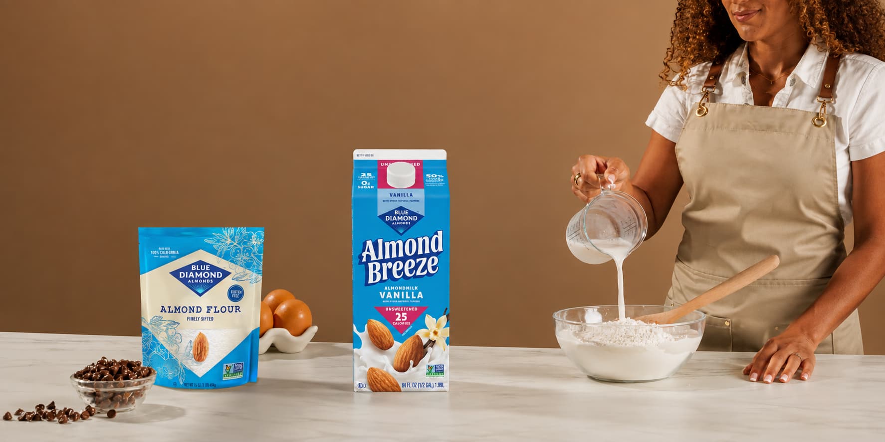A woman pours almond milk into a bowl of flour; almond flour and almond milk products are on the counter with eggs and chocolate chips nearby.