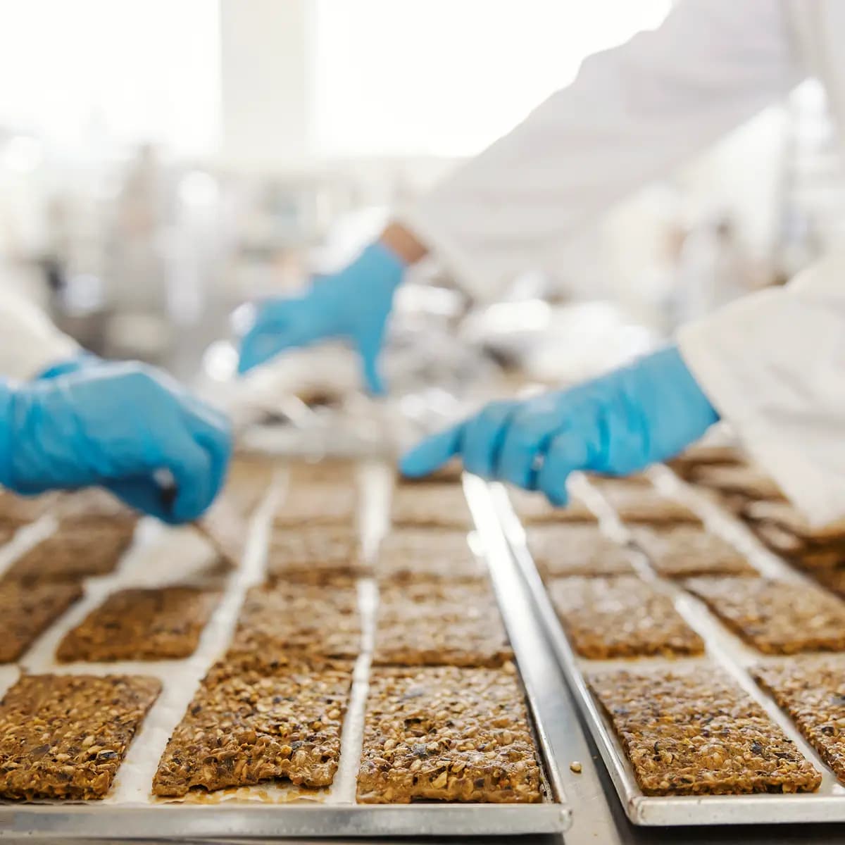 Workers wearing blue gloves arrange rectangular food bars on baking trays in a commercial food production facility.