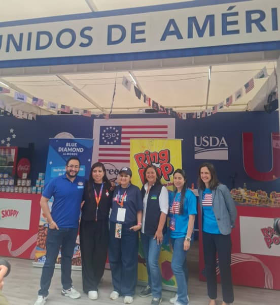 Six people pose together at a USDA booth decorated with U.S. flags and displays for American food brands, including Blue Diamond Almonds and Ring Pop.