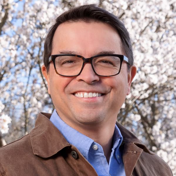 A smiling man wearing glasses, a blue button-down shirt, and a brown jacket, standing in front of blooming white trees.