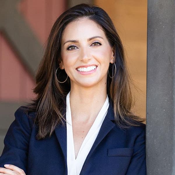 A smiling woman in a navy blazer and white top leaning against a door frame.