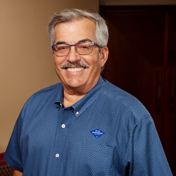 A smiling man with a mustache and glasses wearing a blue short-sleeved button-down shirt with a Blue Diamond Growers logo pinned to the chest.